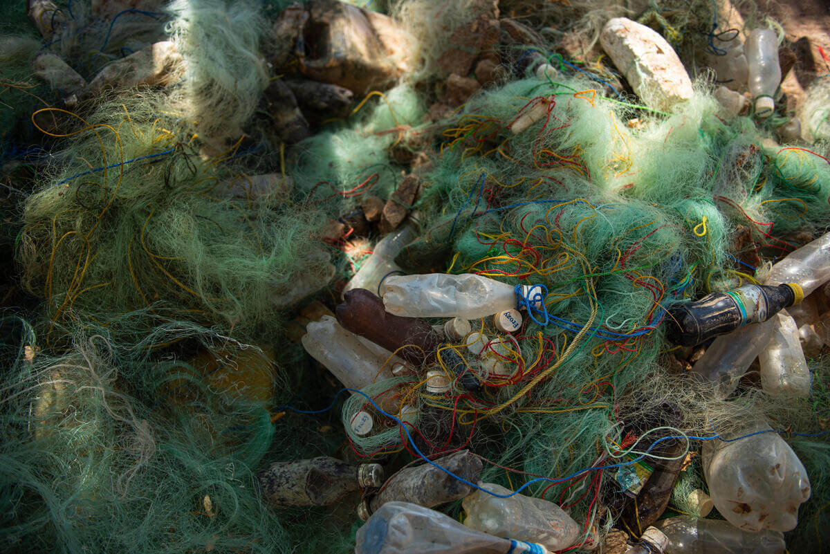 Tangled green fishing nets mixed with various discarded plastic bottles and caps, scattered on a rocky surface. The nets have colorful threads intertwined, and the bottles appear dirty and crushed.
