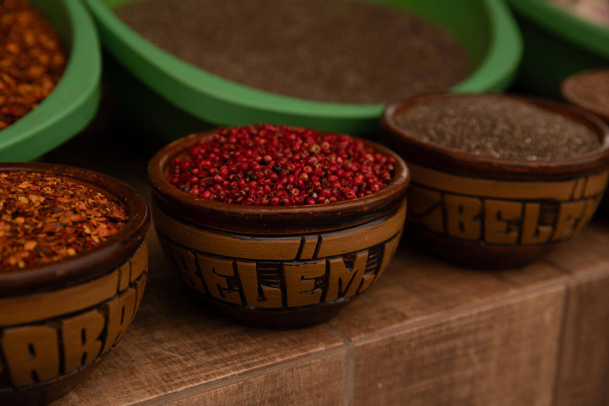 Wooden bowls filled with various spices, including red peppercorns and crushed red chili flakes, arranged on a wooden surface. Green bowls with ground spices are visible in the background.