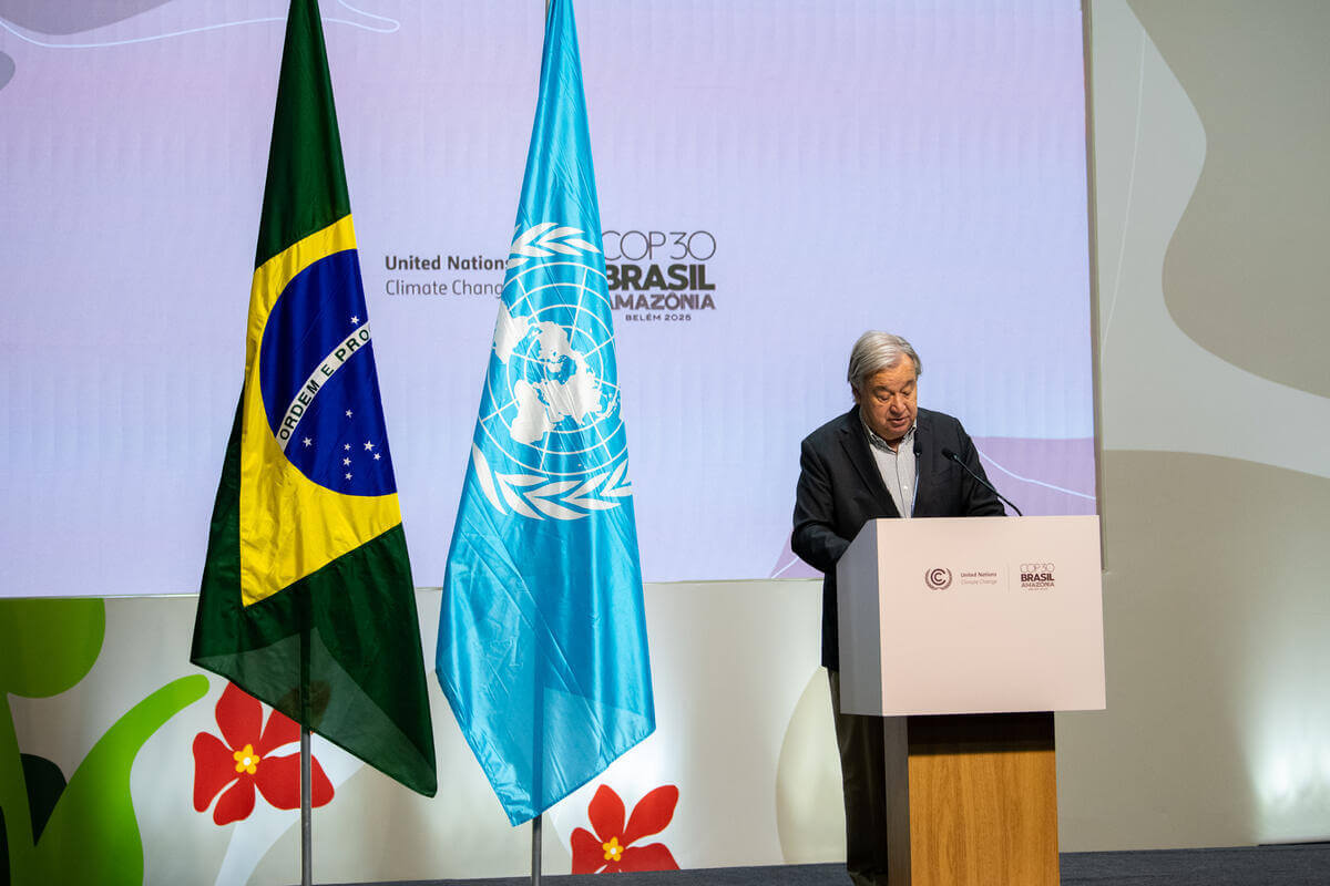 A man in a dark suit jacket and light shirt stands at a white podium with microphones. Behind him are two flags: the Brazilian flag on the left and the United Nations flag on the right. The backdrop displays the text "United Nations Climate Change COP30 Brasil Amazônia Belém 2025.