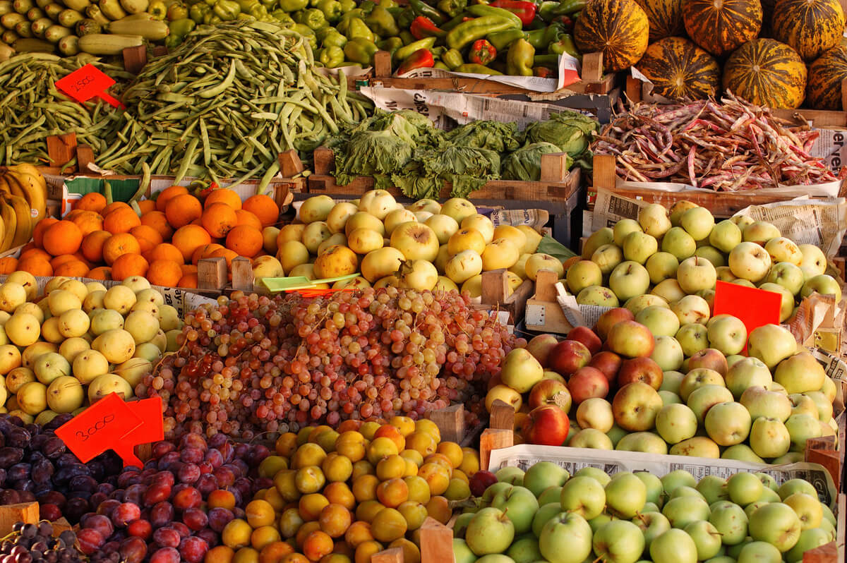 A colorful market display featuring a variety of fresh fruits and vegetables arranged in wooden crates. Visible produce includes green beans, lettuce, green bell peppers, striped melons, oranges, apples in different shades, grapes, plums, bananas, and yellow plums. Bright red price tags are placed among the produce.