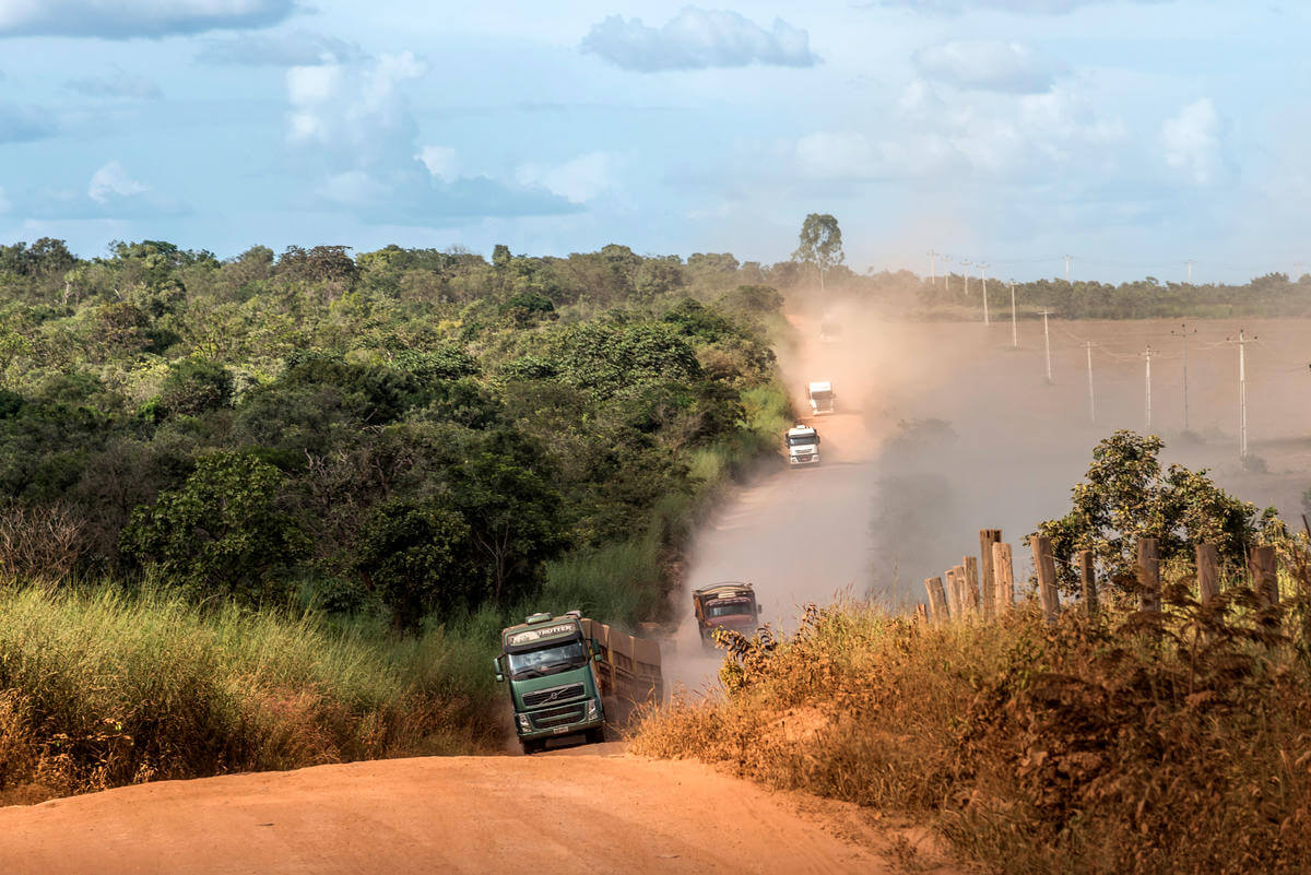 A dusty dirt road cutting through a rural landscape with dense green trees on the left and dry grass with wooden fence posts on the right. Several trucks are driving along the road, kicking up a large cloud of dust behind them. The sky is partly cloudy with patches of blue.