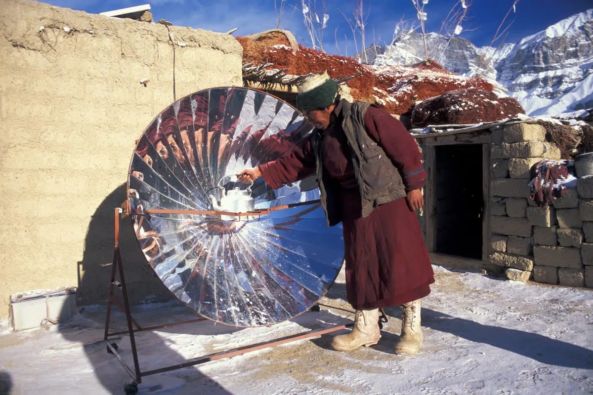 A person wearing a maroon robe, brown vest, green hat, and beige boots is adjusting a white kettle placed on a large parabolic solar cooker with reflective panels. The scene is outdoors on a snowy ground, with a stone and mud wall and a small stone building in the background, set against a backdrop of snow-covered mountains under a clear blue sky.