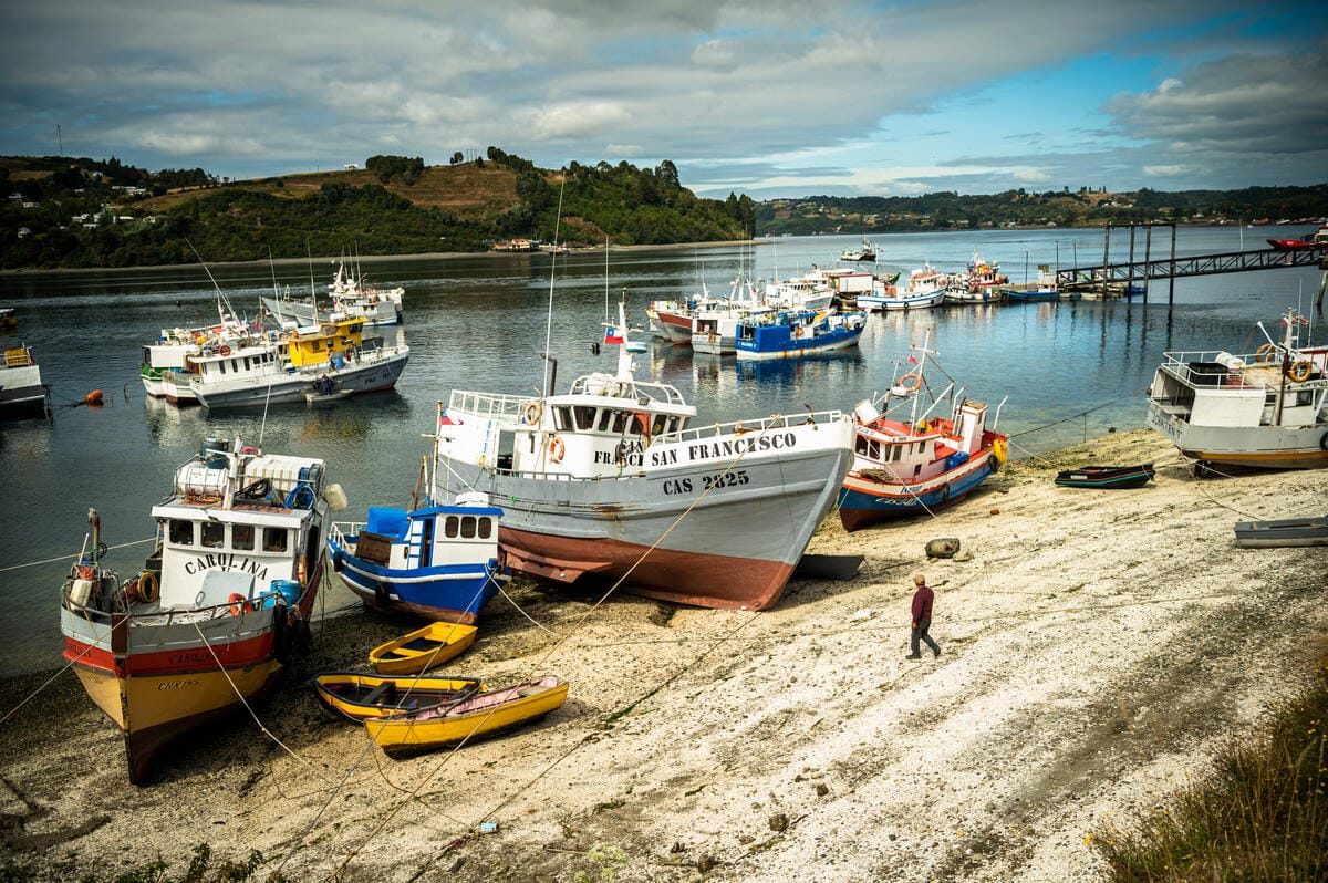 Several fishing boats are docked along a rocky shoreline, with some boats anchored in the water and others resting on the shore. The boats are painted in various colors, including white, blue, yellow, and red. A person is walking on the shore near the boats. In the background, there are green hills and a partly cloudy sky.