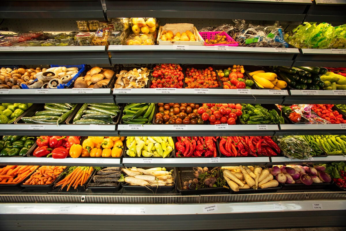 Shelves filled with a variety of fresh vegetables and fruits including mushrooms, tomatoes, cucumbers, peppers, carrots, parsnips, and turnips, all neatly arranged in trays and baskets in a grocery store produce section.