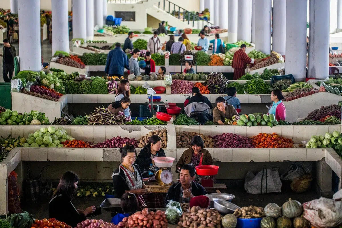 Indoor market with multiple vendors selling a variety of fresh vegetables including cabbages, tomatoes, potatoes, eggplants, and leafy greens. The market features tiled stalls arranged in rows, with several people shopping and working behind the counters. Large white columns support the ceiling, and the atmosphere is busy with activity.