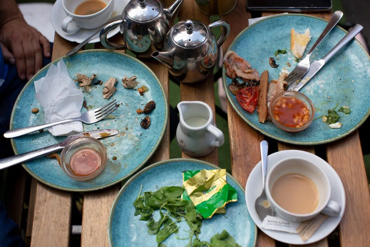 Three blue plates with food remnants, including some greens, beans, a sausage, bacon, and a small container of baked beans. Two cups of tea or coffee, one with milk and a spoon inside, and two stainless steel teapots. A small white milk jug is also on the wooden table. Cutlery and a crumpled napkin are on the plates.