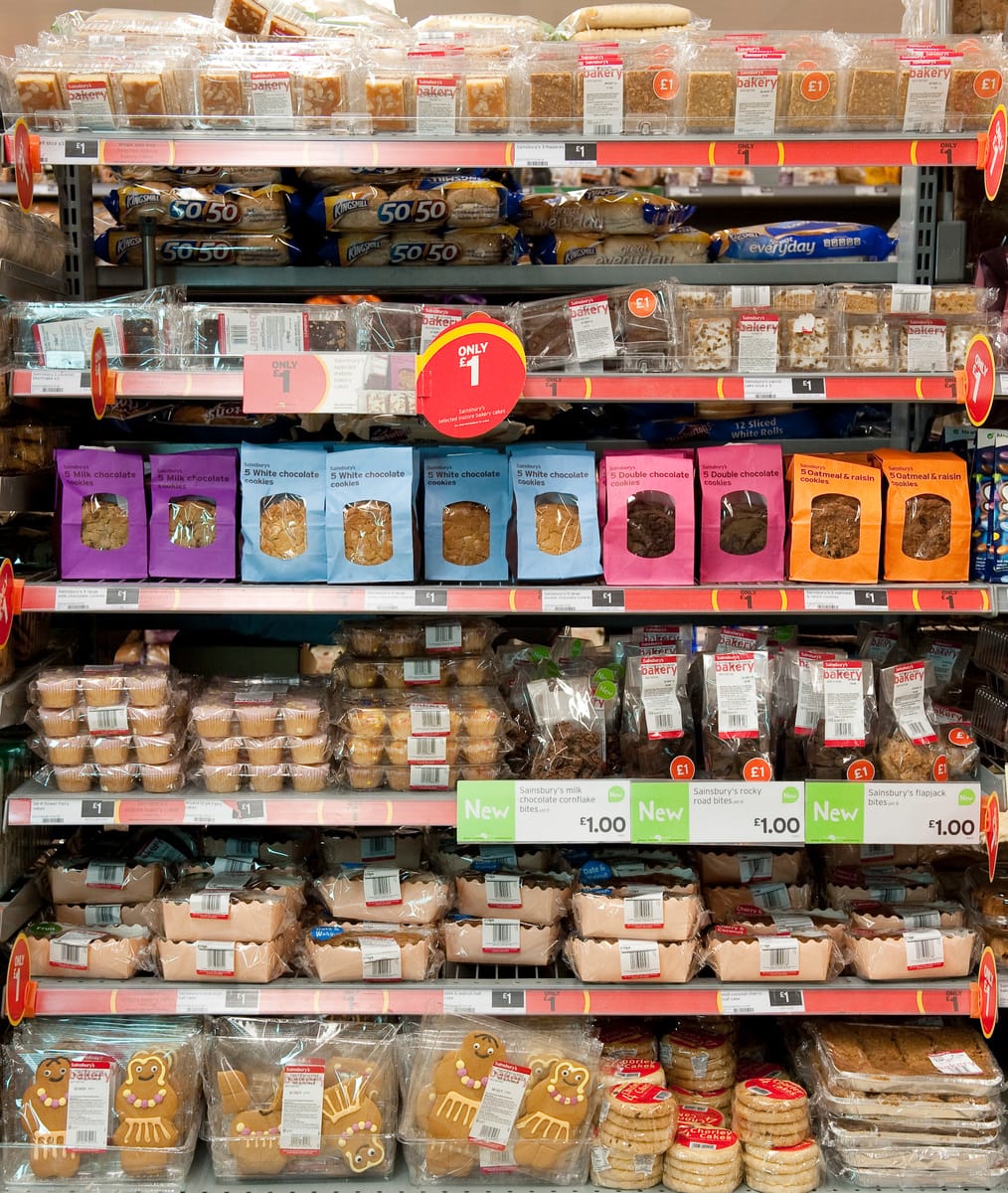 Shelves stocked with various packaged baked goods including cookies, chocolate cookies, oat and raisin cookies, muffins, gingerbread figures, and other assorted bakery items, all labeled with price tags mostly showing £1.00. The packaging features different colors such as purple, blue, pink, orange, and clear plastic. Some products are labeled as new.