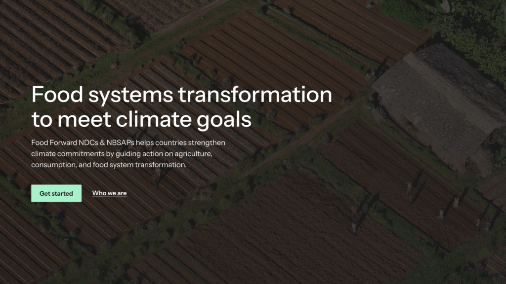 Aerial view of neatly plowed agricultural fields with a small thatched-roof hut on the right side, bordered by green vegetation. The fields are divided into rectangular plots with visible furrows. Several people are scattered around the fields, casting long shadows. Text overlay reads: "Food systems transformation to meet climate goals. Food Forward NDCs & NBSAPs helps countries strengthen climate commitments by guiding action on agriculture, consumption, and food system transformation." There are two buttons below the text labeled "Get started" and "Who we are.